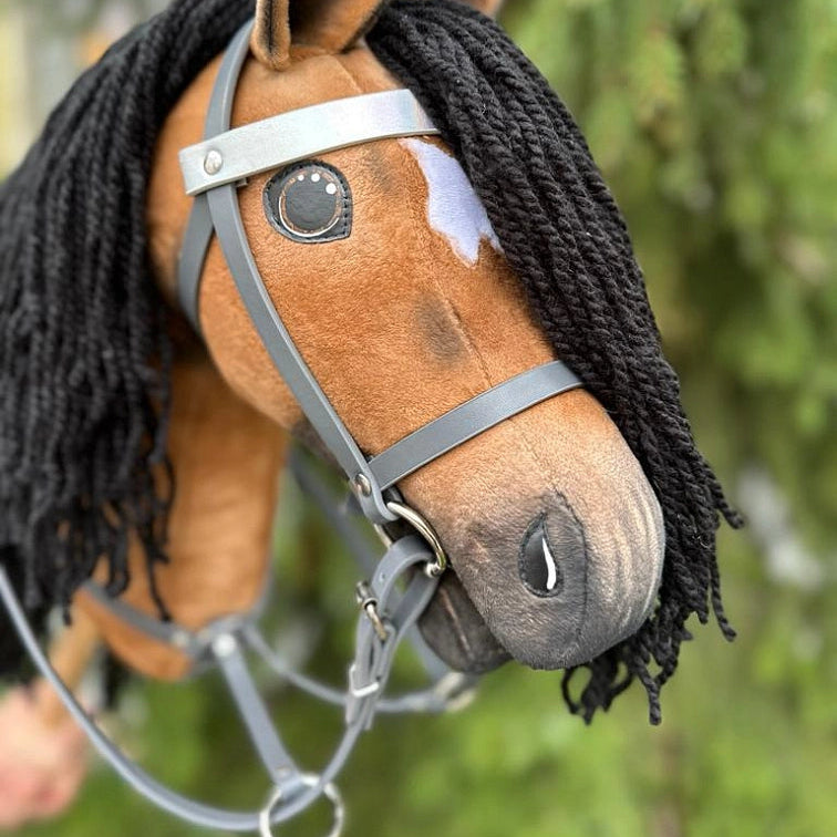 A close-up of a handmade Hobby Horse with a black yarn mane, brown body, a white patch above one eye, and a gray bridle, set against a blurred green outdoor background—a perfect accessory for Hobby Horsing adventures.