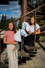 Lifestyle photo: two girls with white Hobby Horses by Hobby Horse LarDen on a rusty metal staircase, the Hobby Horse Lady with Black Halter in front - white plush head, white yarn mane - shown here with the Pastell Blue fur halter in leatherette and the Double Blue knitted lead rope.