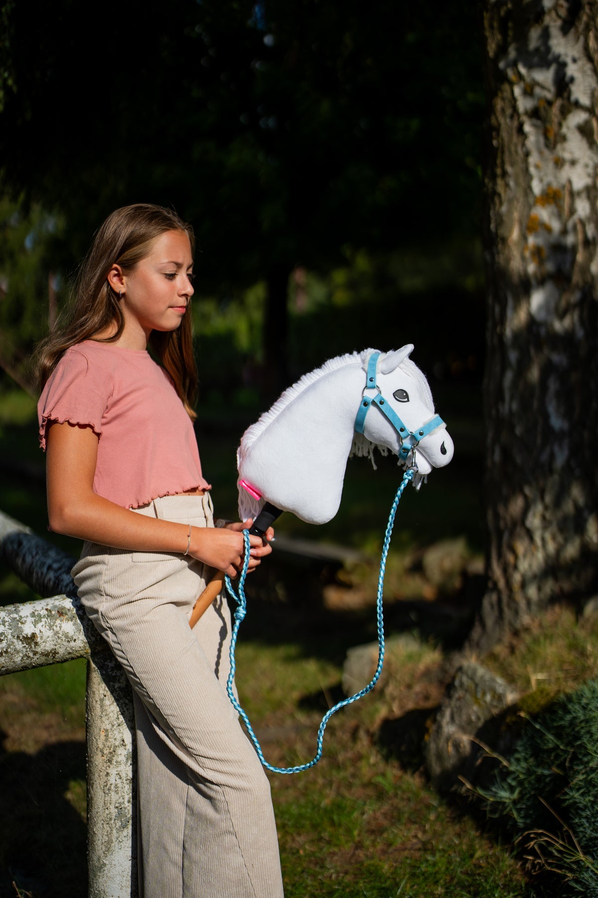 Mädchen im rosa Oberteil steht an einem moosigen Baum und hält das Hobby Horse Lady mit schwarzem Halfter von Hobby Horse LarDen, der Führstrick hängt in einer Schlaufe – weißer Plüschkopf, weiße Garnmähne – hier mit dem Pastell Blue Fellhalfter aus Kunstleder und dem gestrickten Double Blue Führstrick