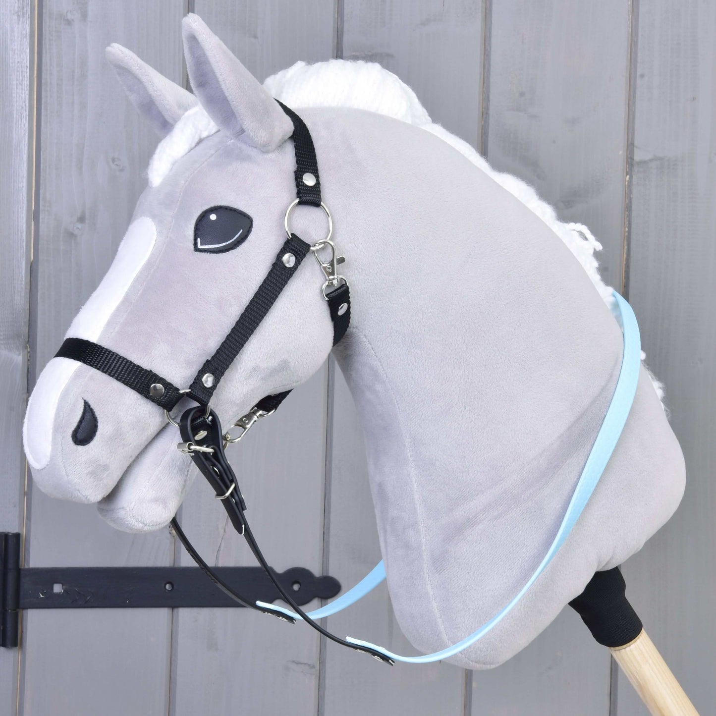 Graues Plüsch-Hobby Horse mit weißer Mähne und Gummierte Zügel - Light blue von Hobby Horse LarDen, befestigt an einem Holzstab, vor einer grauen Holzwand.