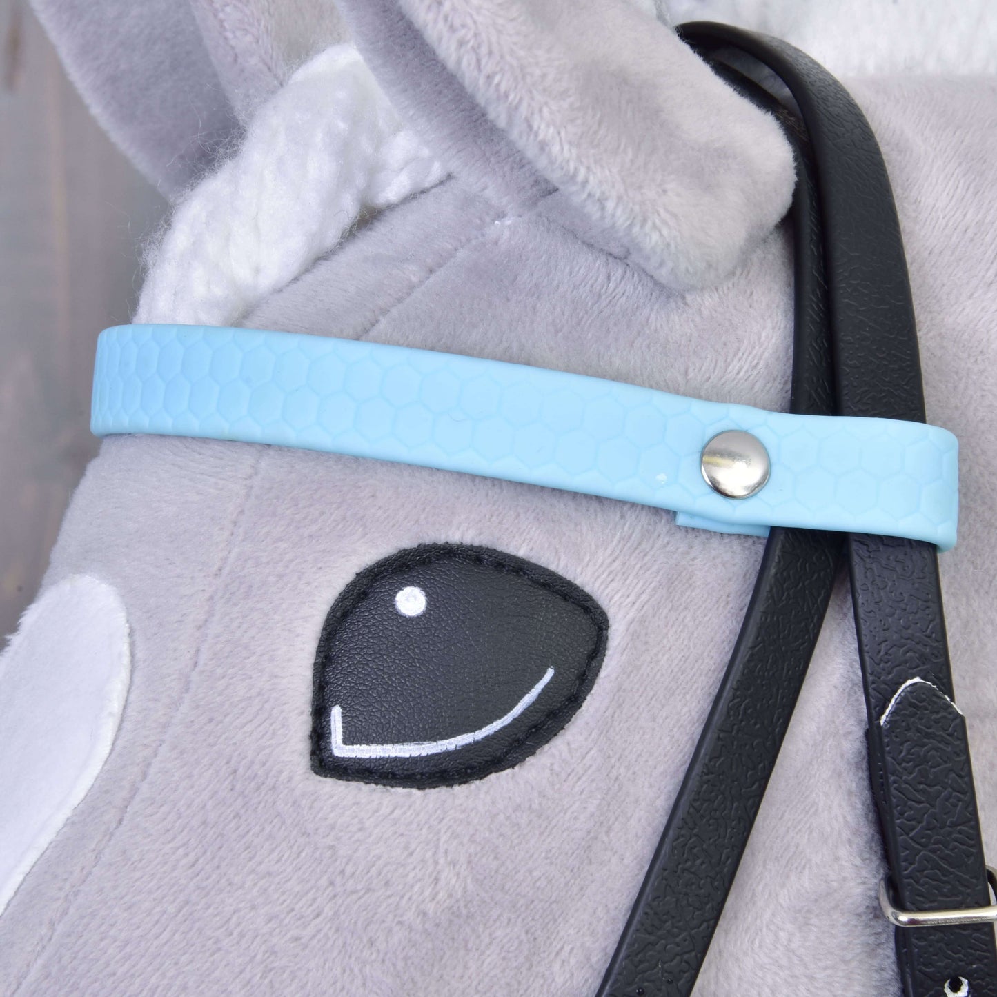 Nahaufnahme des Gesichts eines grauen Plüsch-Hobby Horse mit gesticktem schwarzem Auge, weißem Ohr und dem Komplettes Zaumzeug ohne Nasenriemen Light Blue von Hobby Horse LarDen mit hellblauem Riemen, silberfarbenem Knopf und abnehmbaren schwarzen Zügeln.