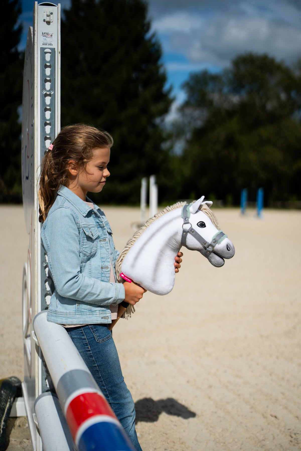 Mädchen in Jeansjacke steht an einem Sprungständer auf einem Sandplatz und hält das Hobby Horse Little Lady mit schwarzem Halfter von Hobby Horse LarDen – weißer Plüschkopf, cremefarbene Garnmähne – hier mit dem grauen Halfter mit mintfarbenem Fell