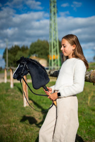 Lifestyle photo: a girl in a white jumper stands in a sunlit meadow holding the Hobby Horse Loki with Black Halter by Hobby Horse LarDen - black plush head, white blaze, black yarn mane - shown here with the complete Pure Black bridle without noseband.