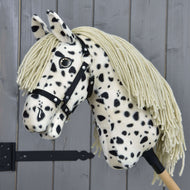 Hobby Horse Lord Valentine mit Schwarzem Halfter von Hobby Horse LarDen mit geflecktem Plüschfell, langer cremefarbener Mähne, Knopfauge und auf einem Holzstock vor grauen Holzpaneelen.