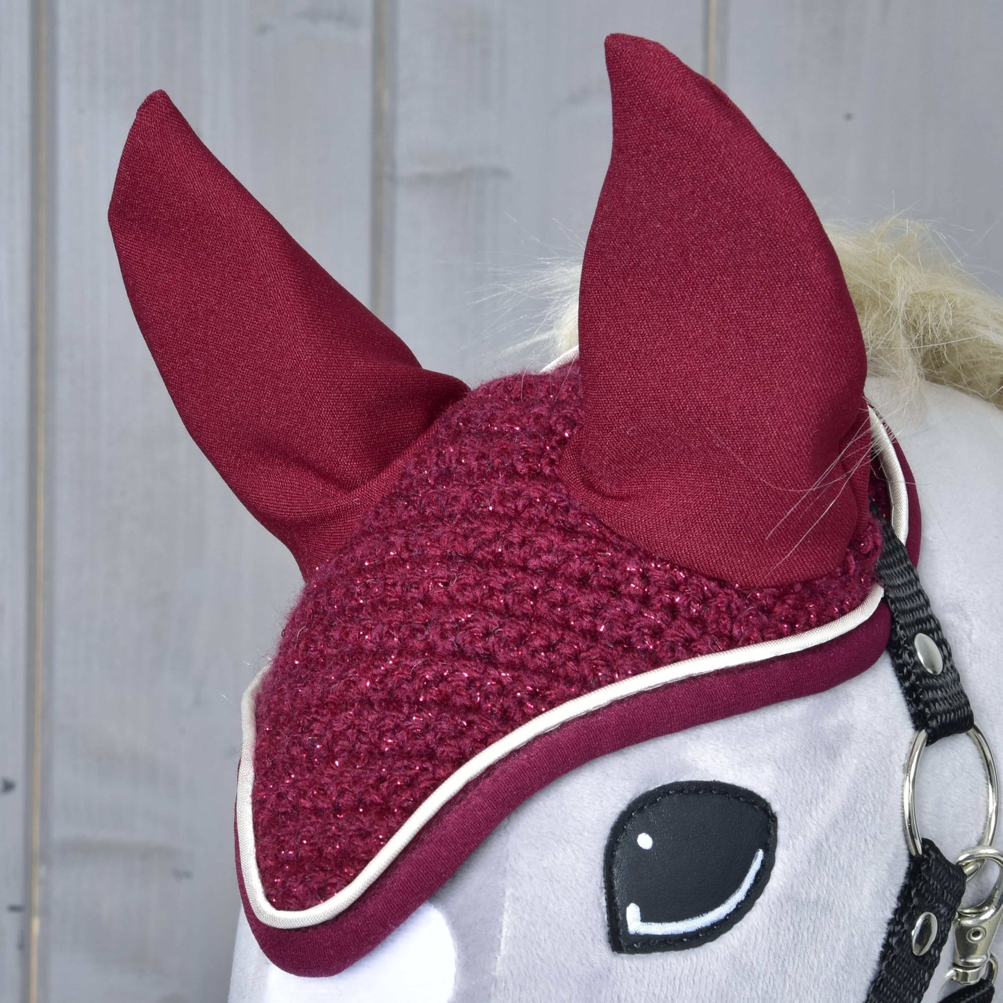 Nahaufnahme der Ohrenkappe gehäkelt Lux Maroon von Hobby Horse LarDen mit hohen, spitzen Ohren und weißem Rand auf einem weißen Hobby Horse vor Holz-Hintergrund.