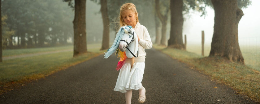 A young girl in a white dress walks down a tree-lined, foggy road, clutching her colorful Hobby Horse.