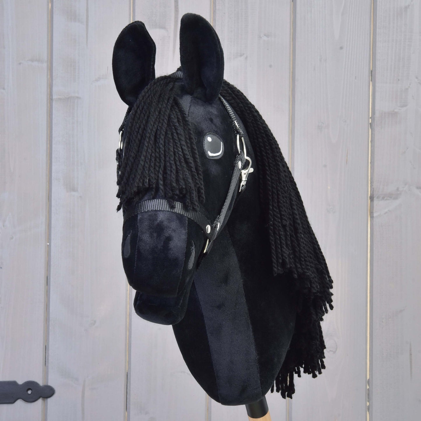 Hobby Horse Mahal avec licol noir de Hobby Horse LarDen, avec tête noire en peluche inspirée du frison, crinière en fil et bride, monté sur un bâton et présenté sur fond en bois clair.