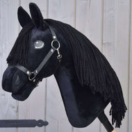 Hobby Horse Mahal mit Schwarzem Halfter von Hobby Horse LarDen, ein Hobby Horse im Friesen-Stil aus schwarzem Plüsch mit schwarzer Mähne, schwarzem Halfter und weißem Augendetail, auf einem Holzstab montiert und vor einer hellgrauen Holzwand gezeigt.