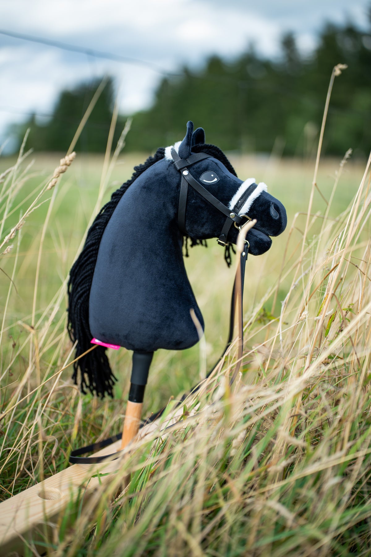 Hobby Horse Mahal mit schwarzem Halfter von Hobby Horse LarDen steht in hohem trockenem Gras auf einer Wiese – schwarzer Plüschkopf, schwarze Garnmähne, Holzstab – mit vollständigem Zaumzeug mit Fell