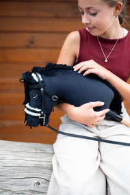 Lifestyle photo: a girl in a burgundy top holds the Hobby Horse Mahal with Black Halter by Hobby Horse LarDen in her arms in front of a wooden wall - black plush head, black yarn mane, white fur noseband - shown here with the complete bridle with fur.