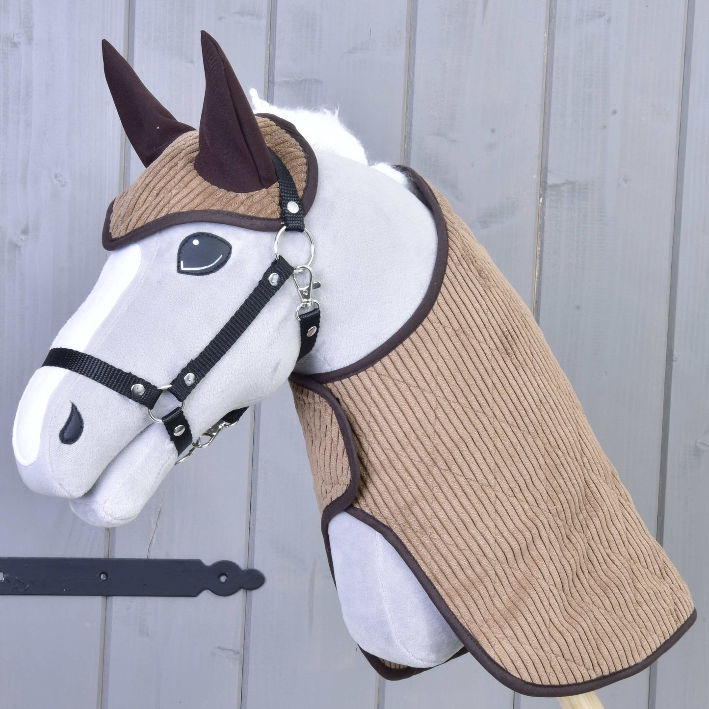 Das Set Manchester Le Chocolate von Hobby Horse LarDen zeigt ein Plüsch-Hobby Horse mit weißem Gesicht, schwarzem Zaumzeug, braunen Ohren und strukturiertem braunem Stoff an Kopf und Hals, mit einer Hobby Horse Decke auf einem Holzstab vor einer grauen Holzwand.