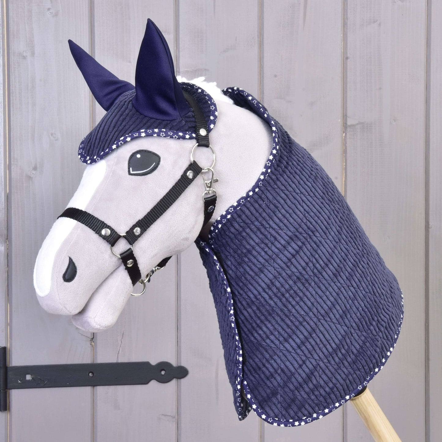 Das Hobby Horse LarDen Set Manchester Blue Star zeigt ein graues Hobby Horse mit schwarzem Zaumzeug und Zügeln sowie einer navyblauen gesteppten Decke und Fliegenhaube mit weißer Einfassung, auf einem Holzstab vor hell getäfeltem Holz-Hintergrund.
