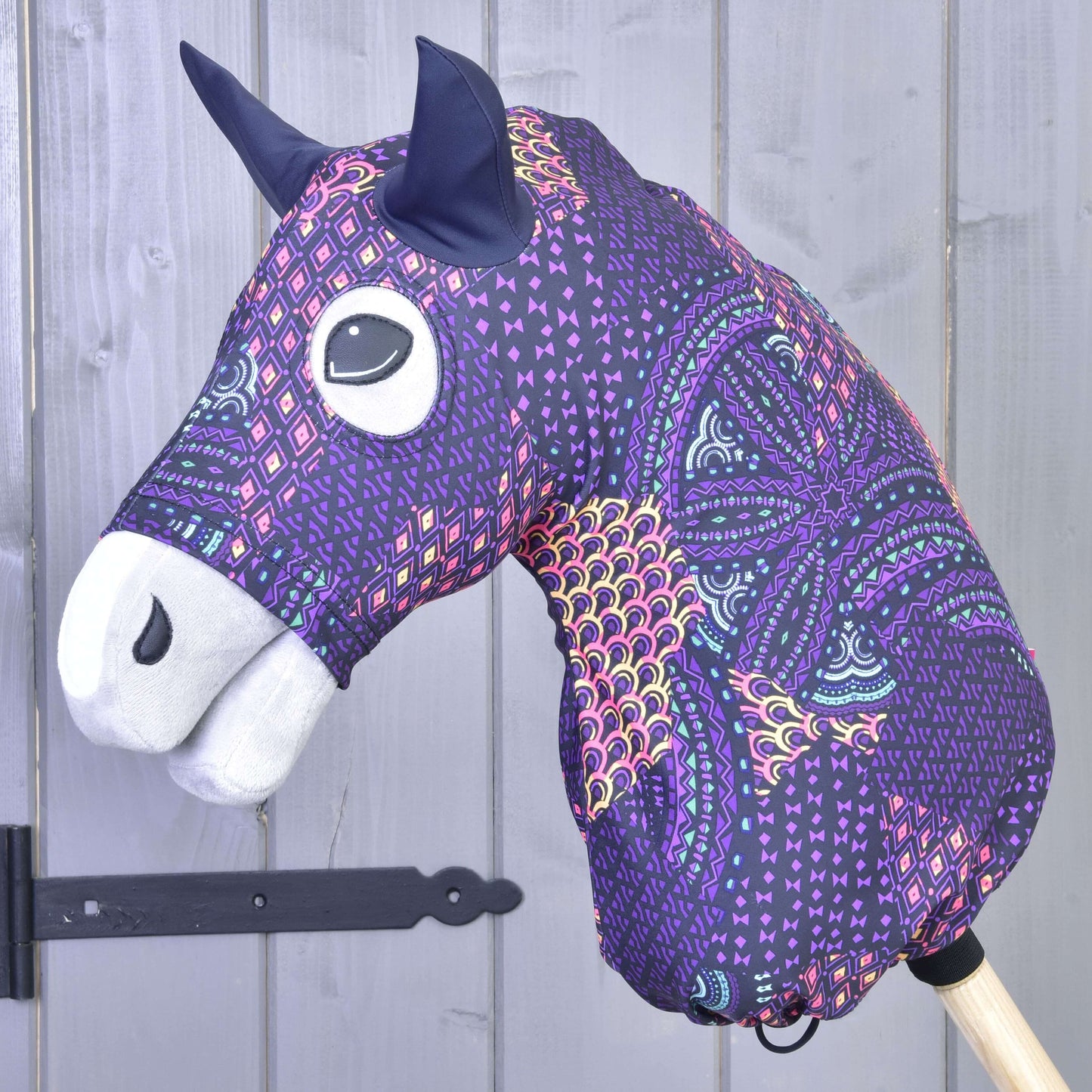 Hobby Horse Schutzanzug Mandala Purple  (Lycra) von Hobby Horse LarDen mit Stoffkopf in lila, pinken und orangefarbenen geometrischen Mustern, schwarzen Ohren und weißer Schnauze, zusammen mit einem stilvollen Accessoire vor grauem Holzhintergrund gezeigt.