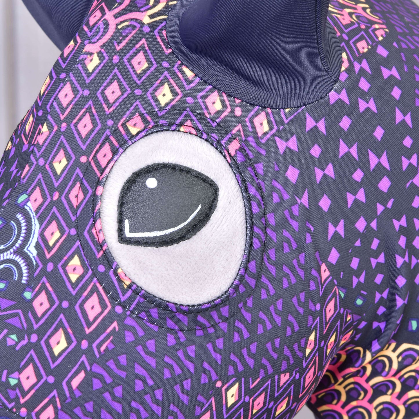 Nahaufnahme des Hobby Horse Schutzanzug Mandala Purple  (Lycra) von Hobby Horse LarDen mit großem gesticktem Auge und lila Stoff mit bunten geometrischen und Rautenmustern; oben ist ein dunkles Ohr zu sehen.