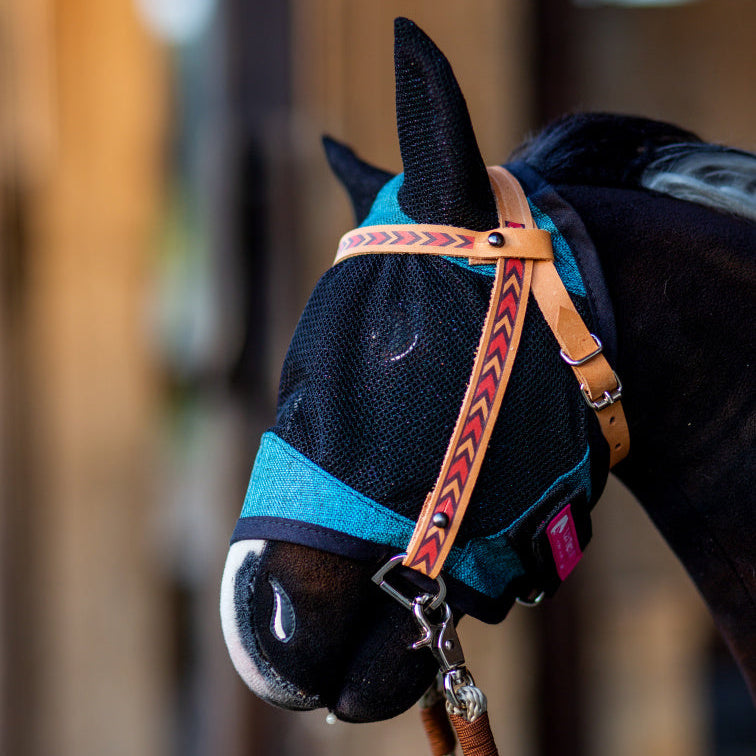 Nahaufnahme eines Pferdes mit blauer Fliegenmaske aus Mesh mit Ohrenschutz und einem braunen Lederhalfter mit rotem Pfeilmuster, das an Ausrüstung für Hobby Horsing erinnert. Der Hintergrund ist weich unscharf.