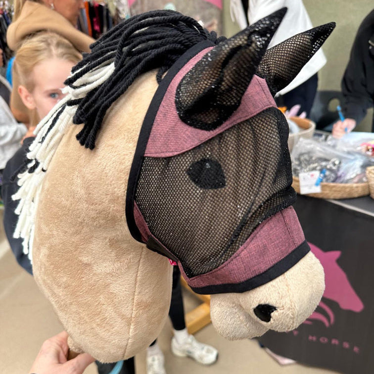 Nahaufnahme eines handgefertigten Hobby Horse mit Mesh-Maske und schwarz-weißer Garnmähne. Im unscharfen Hintergrund zeigen Menschen und ein Stand mit pferdebezogenen Artikeln den kreativen Geist des Hobby Horsing.