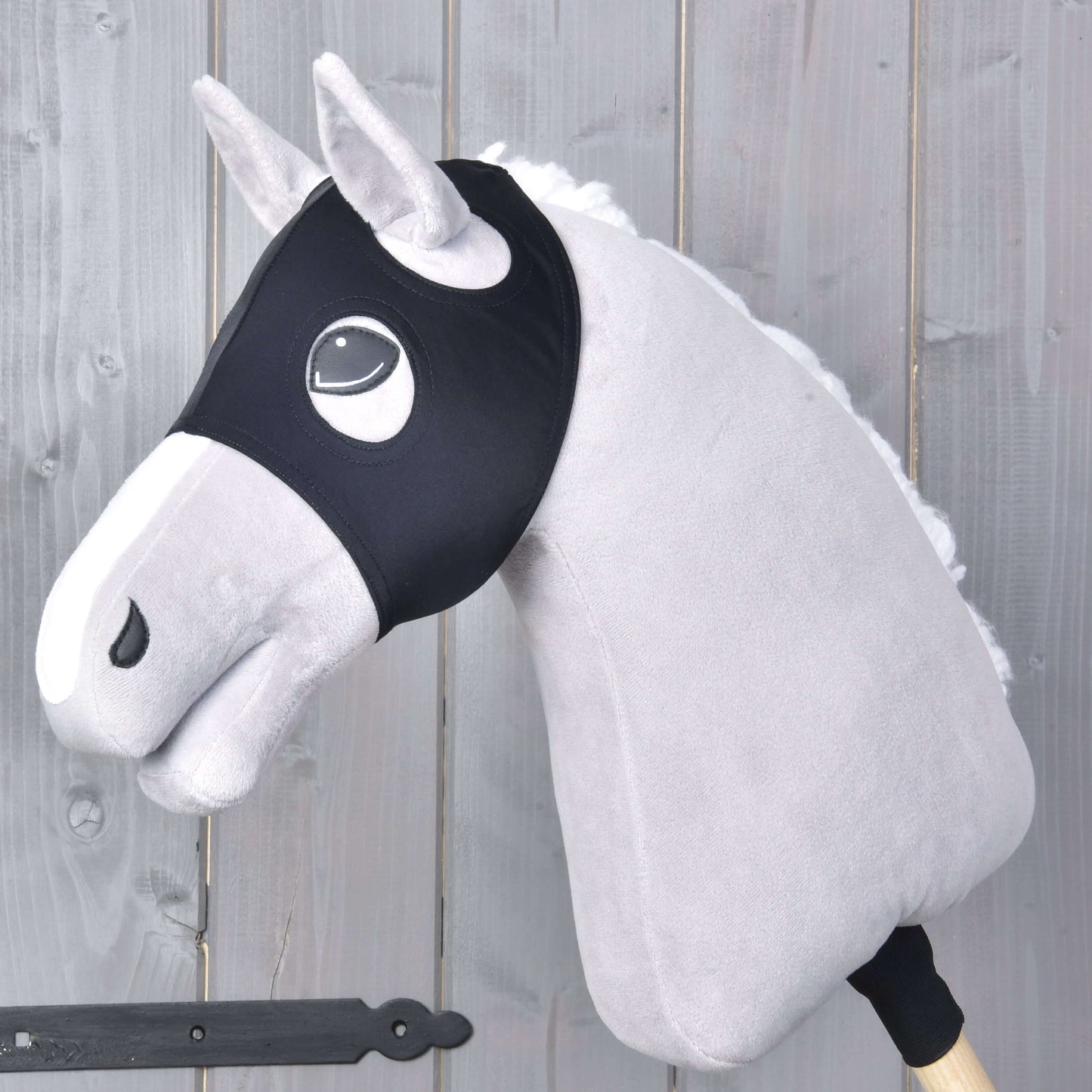 Ein Plüsch-Hobby Horse mit hellgrauem Fell, weißer Mähne und schwarzem Zaumzeug trägt Maske ohne Ohren schwarz von Hobby Horse LarDen und ist auf einem Holzstab vor einem Hintergrund aus grauen Holzpaneelen montiert.