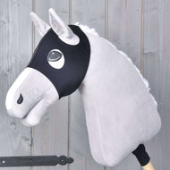 Ein Plüsch-Hobby Horse mit hellgrauem Fell, weißer Mähne und schwarzem Zaumzeug trägt Maske ohne Ohren schwarz von Hobby Horse LarDen und ist auf einem Holzstab vor einem Hintergrund aus grauen Holzpaneelen montiert.