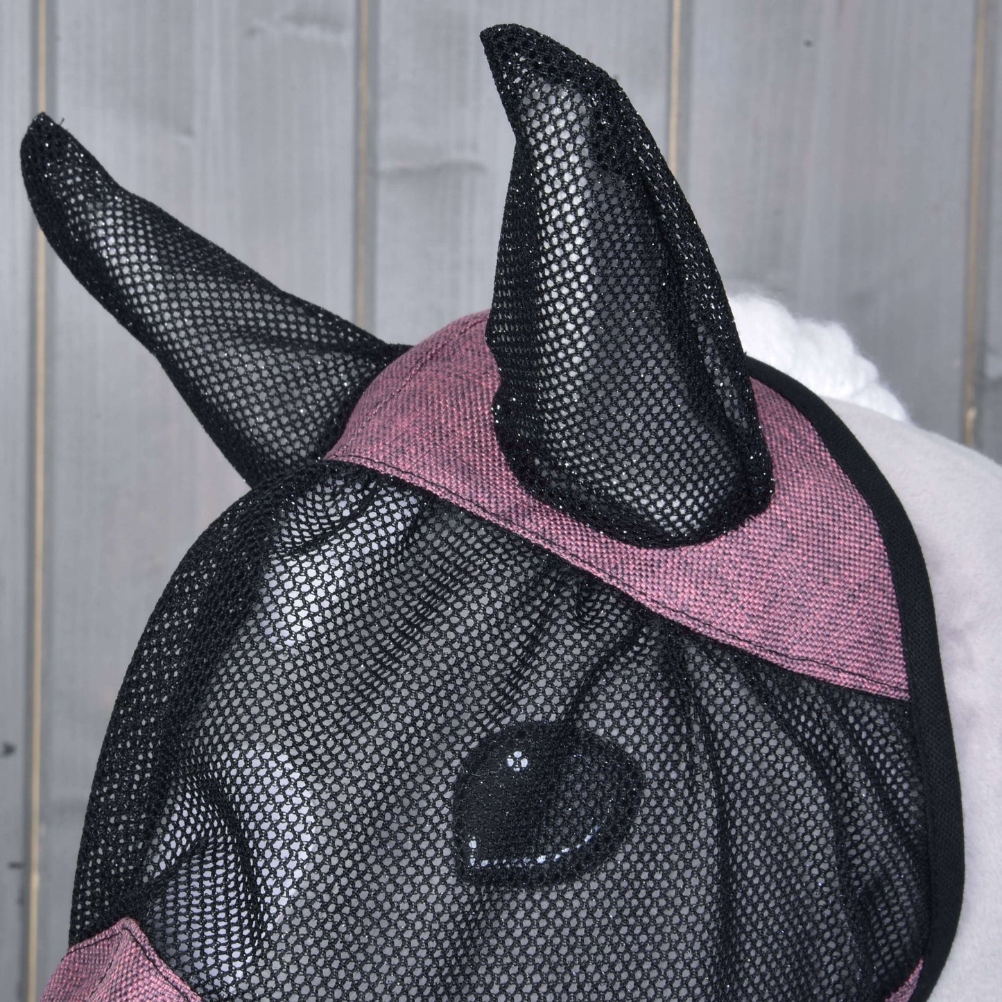 Nahaufnahme eines Pferdes mit der Fliegenmaske de Luxe Pink von Hobby Horse LarDen, mit kastanienroter Einfassung und Bedeckung von Ohren, Augen und einem Teil des Gesichts für vollständigen Schutz. Vor hellem Holz Hintergrund.