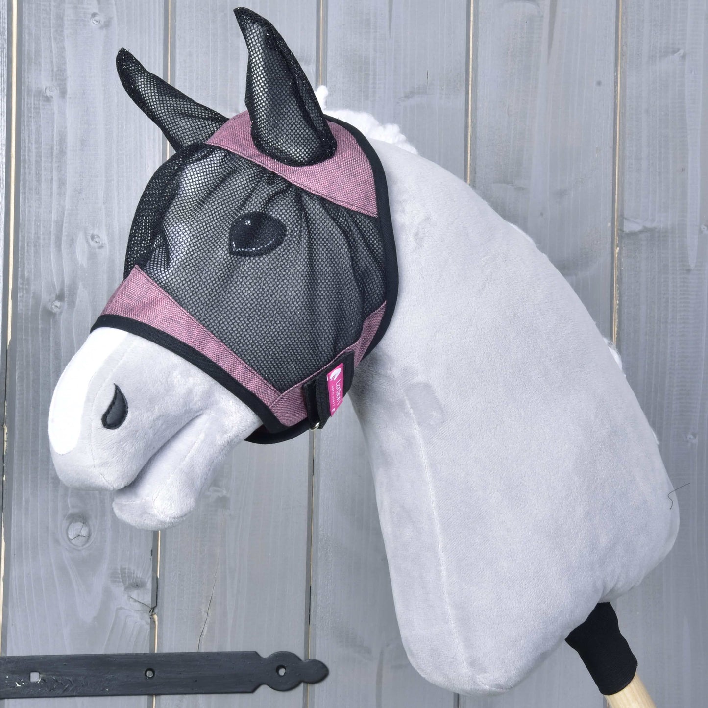 Die Hobby Horse LarDen Fliegenmaske de Luxe Pink, ein graues Stoff-Hobby Horse mit rosa Besatz und waschbarer Fliegenmaske, ist auf einem Holzstab montiert und vor einem Hintergrund aus Holzplanken zu sehen.