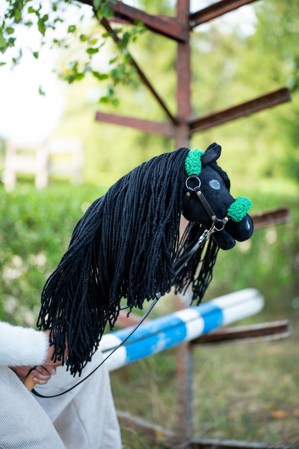 Detail photo: the head and long mane of the Hobby Horse Maximus with Black Halter by Hobby Horse LarDen held up by a wooden jump, greenery behind - black plush, black yarn mane - shown here with the black leatherette halter with green fur and the black leatherette reins.