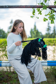 Lifestyle photo: a girl in a pale jumper sits on a paddock rail gathering the reins of the Hobby Horse Maximus with Black Halter by Hobby Horse LarDen - black plush head, long black yarn mane - shown here with the black leatherette halter with green fur and the black leatherette reins.