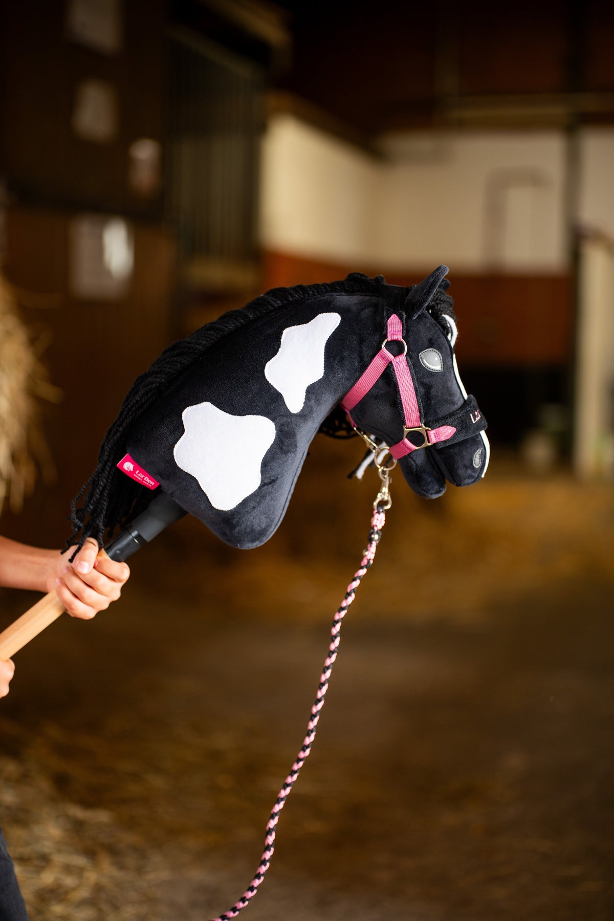 Kopf und Hals des im Stall hochgehaltenen Hobby Horse Merlin mit schwarzem Halfter von Hobby Horse LarDen – schwarzer Plüsch mit weißen Flecken, schwarze Garnmähne – hier mit dem Set LarDen Exclusive Pink und seinem geflochtenen rosa Führstrick gezeigt