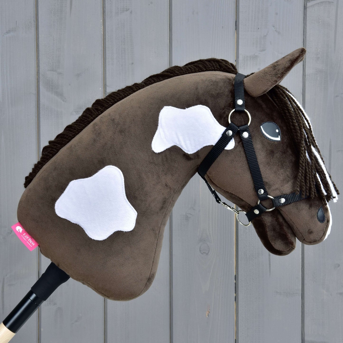 Hobby Horse Monte mit Schwarzem Halfter von Hobby Horse LarDen, ein braunes Plüsch-Hobby Horse mit weißen Flecken und schwarzem Halfter, auf einem Holzstab montiert und vor einem grauen Hintergrund aus Holzplanken gezeigt.