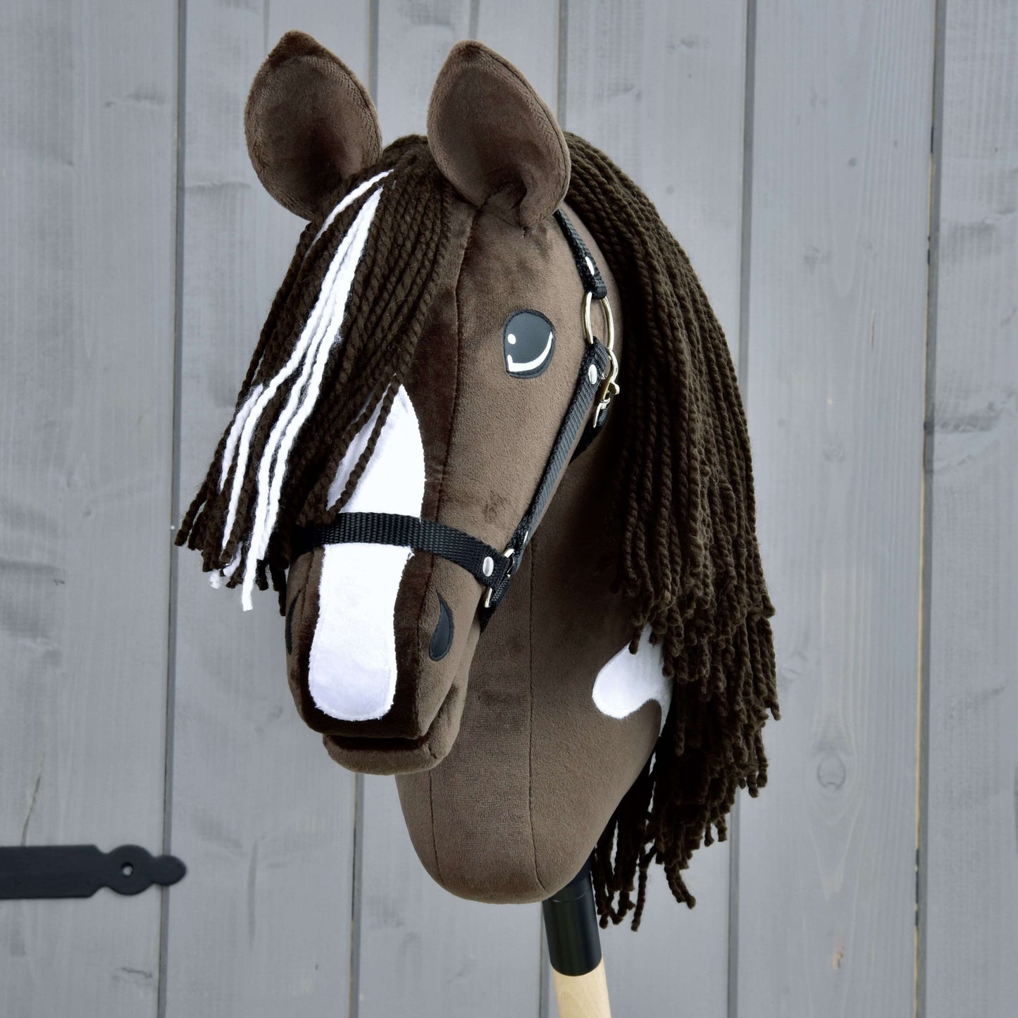 Hobby Horse Monte mit Schwarzem Halfter von Hobby Horse LarDen, ein handgefertigtes Hobby Horse mit braun-weißem Fell im Stil des American Paint Horse, schwarzem Halfter und langer Garnmähne, auf einem Holzstab montiert vor grauem Holz-Hintergrund.