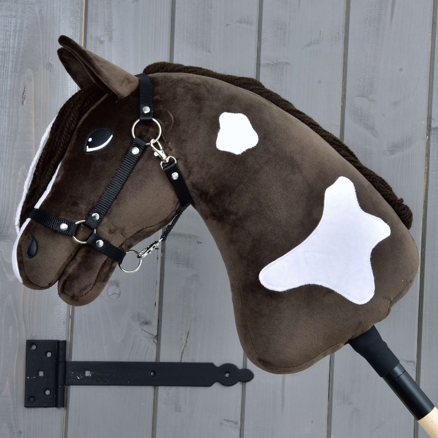 Hobby Horse LarDen „Hobby Horse Monte mit Schwarzem Halfter“ ist ein braun-weißes Plüsch-Hobby Horse, inspiriert vom American Paint Horse, mit freundlichem Gesicht und weißen Flecken, montiert an einer grauen Holzwand.