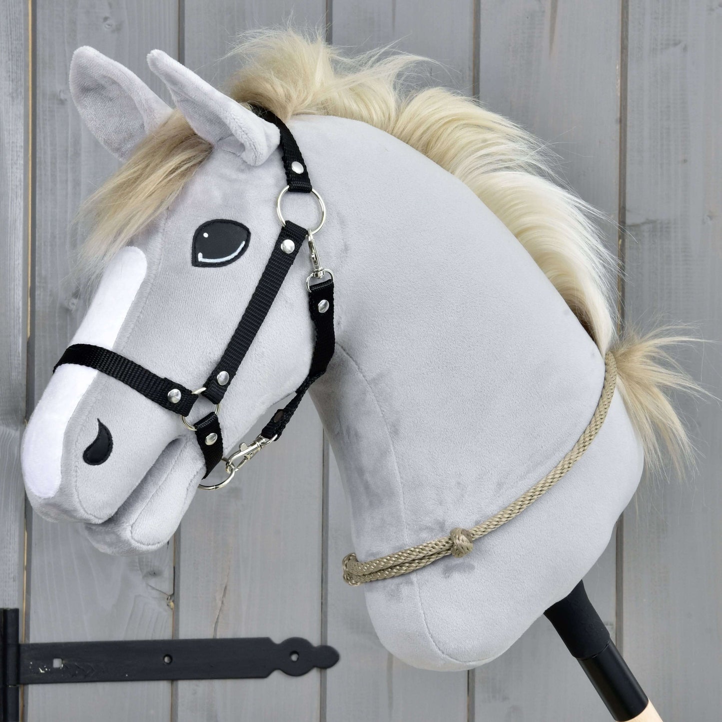 Der Hobby Horse LarDen Halsring Eleganza mit einem grauen Plüschkopf mit schwarzem Zaumzeug und beiger Mähne auf einem Stab, vor Holzplanken – ideal als Übung für das Reiten ohne Zaumzeug oder Cordero-Techniken.