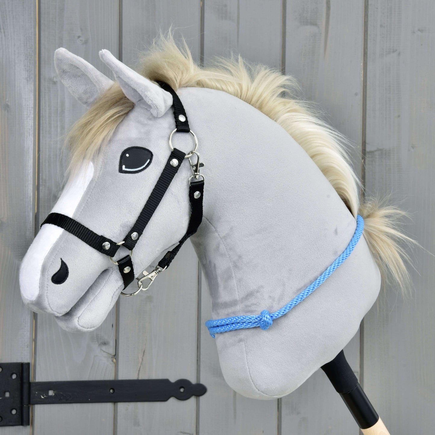 Cordéo Ciel de Hobby Horse LarDen, Hobby Horse en peluche gris avec crinière blanche, bride noire et corde bleue, conçu pour être monté avec ou sans bride. Il se tient devant un fond en bois et affiche une expression amicale.