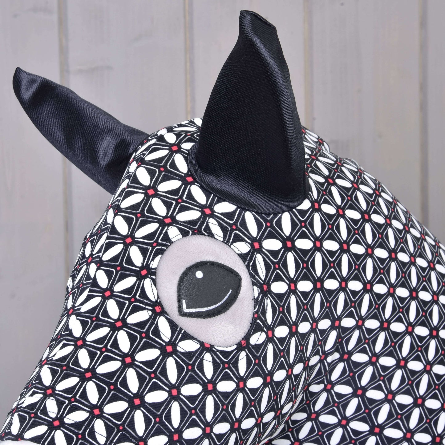 Nahaufnahme des Hobby Horse LarDen Hobby Horse Schutzanzug DeLuxe (Lycra) mit waschbarem schwarzem und weißem geometrischem Lycra-Stoff, großen schwarzen Ohren und gesticktem Auge vor hellem Holz-Hintergrund.