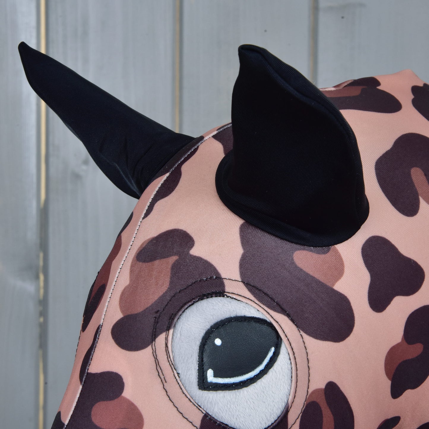 Gros plan sur la combinaison de protection Hobby Horse Glamour de Hobby Horse LarDen, avec tissu à imprimé léopard, grand œil cousu et deux oreilles noires pointues, ressemblant à un Hobby Horse ludique sur fond en bois clair.