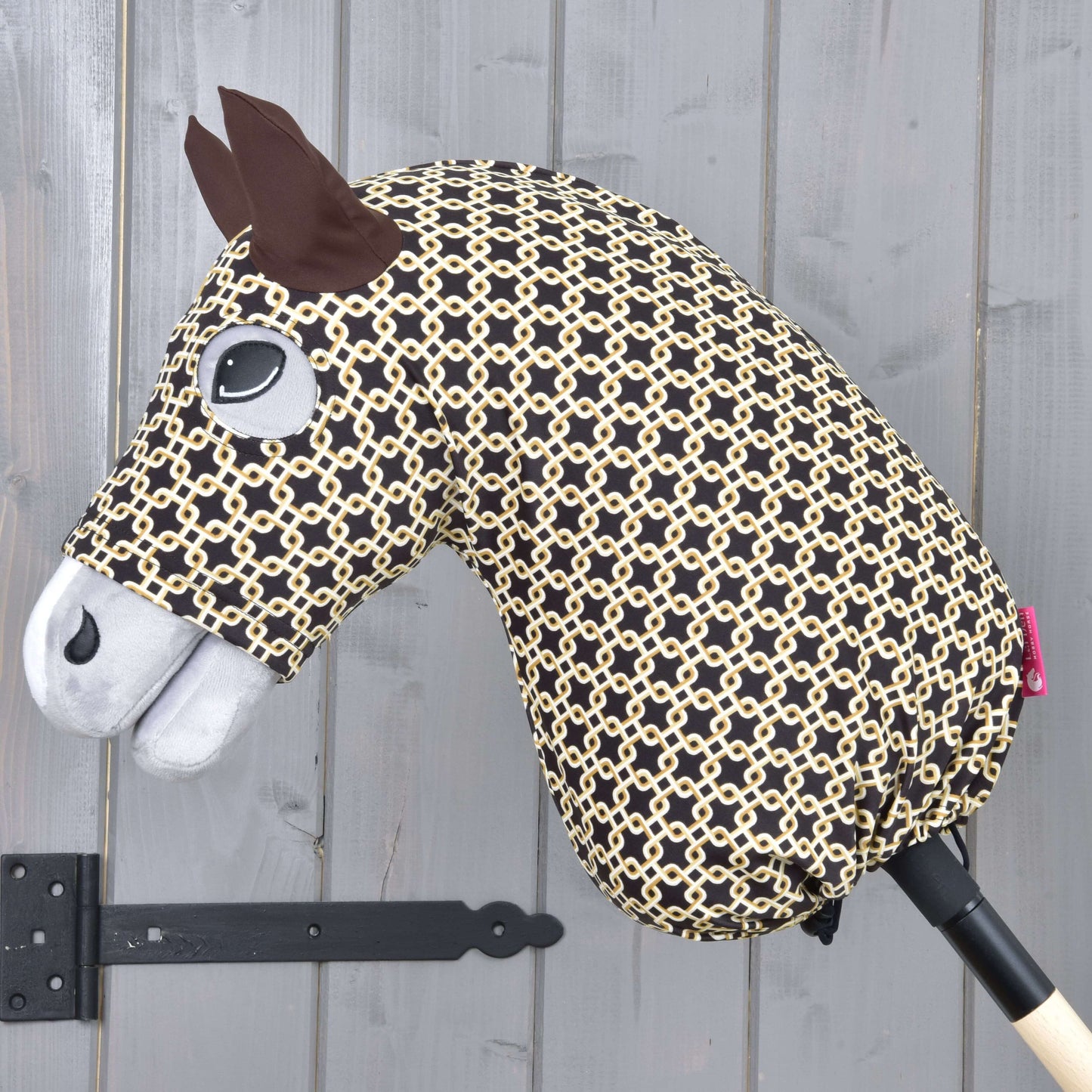 Combinaison de protection Hobby Horse Design Luxe de Hobby Horse LarDen, avec tête noire et beige à motif, oreilles en feutre, œil cousu, museau blanc et matériaux lavables, le tout monté sur un bâton en bois devant un mur gris.
