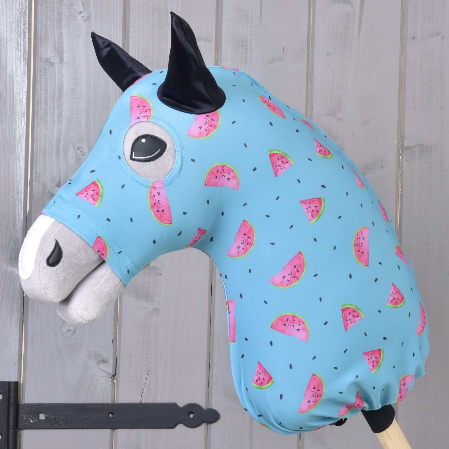 Combinaison de protection Hobby Horse Melon (Lycra) de Hobby Horse LarDen avec housse lavable bleue ornée de tranches de pastèque roses et de graines noires, conçue pour les têtes de Hobby Horse en peluche. Idéale comme protection, présentée sur un bâton en bois devant un mur à panneaux de bois clair.