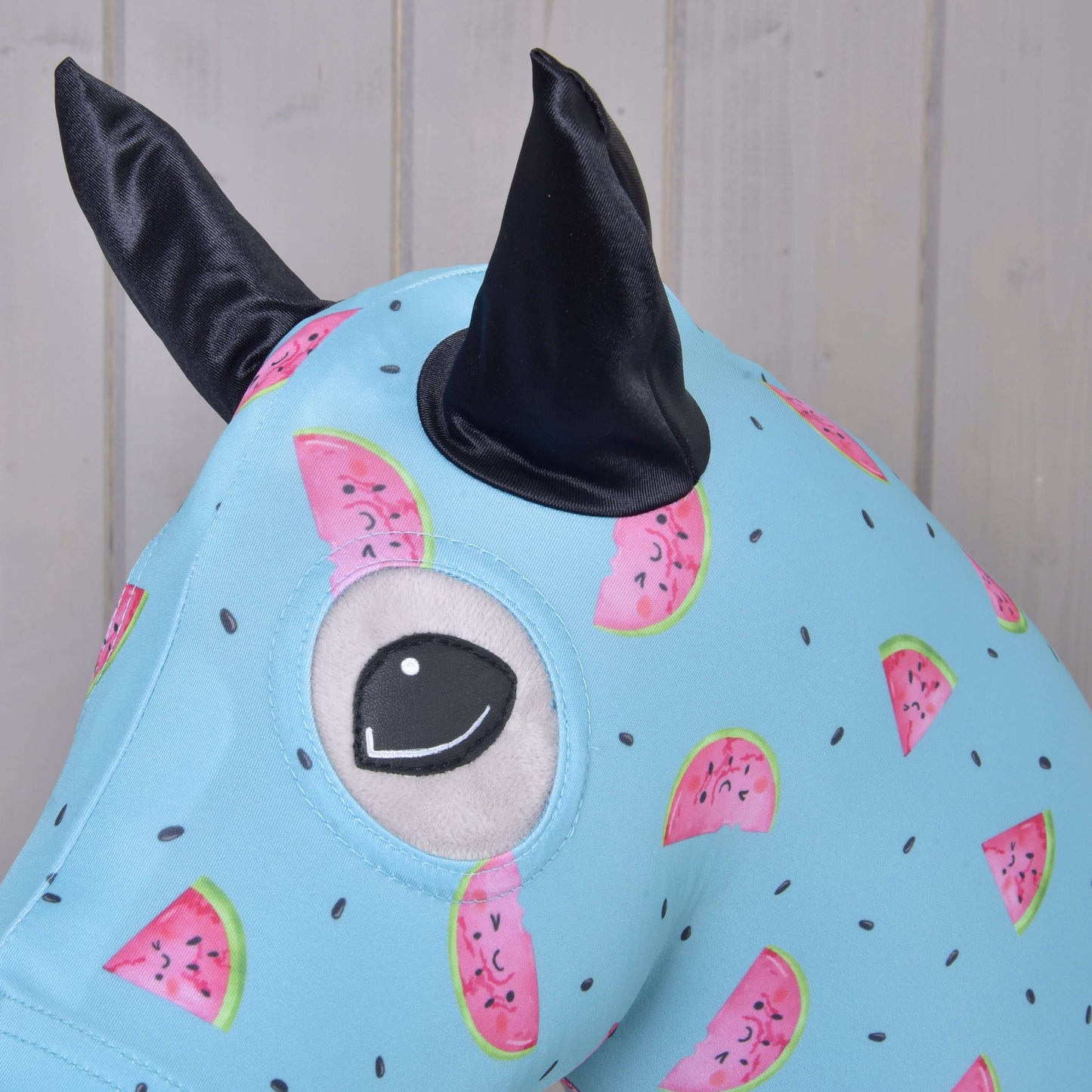 Gros plan sur la Combinaison de protection Hobby Horse Melon (Lycra) de Hobby Horse LarDen, une housse lavable en lycra bleu clair à motif pastèque, avec un grand œil brodé et deux oreilles noires en tissu, sur fond en bois clair.
