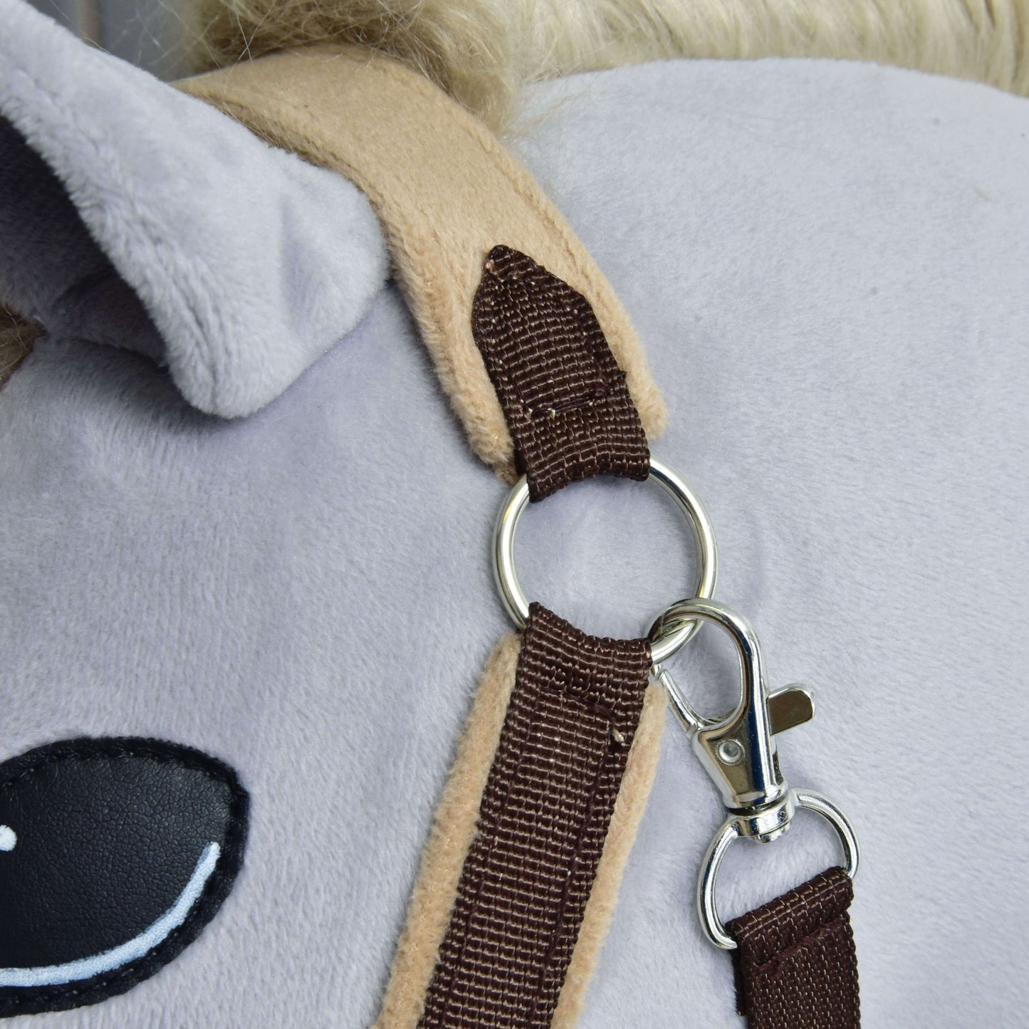 Gros plan sur un cheval en peluche portant le Licol en fourrure LarDen marron de Hobby Horse LarDen, avec muserolle douce en polaire et anneau métallique avec clip près de son oreille et de son œil. La peluche a une surface gris clair et une crinière couleur crème.