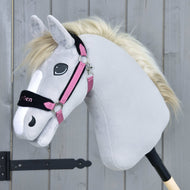 Ein graues Plüsch-Hobby Horse LarDen mit weißer Mähne trägt das Fellhalfter LarDen Pink und sitzt auf einem Stab vor einem Holzzaun.