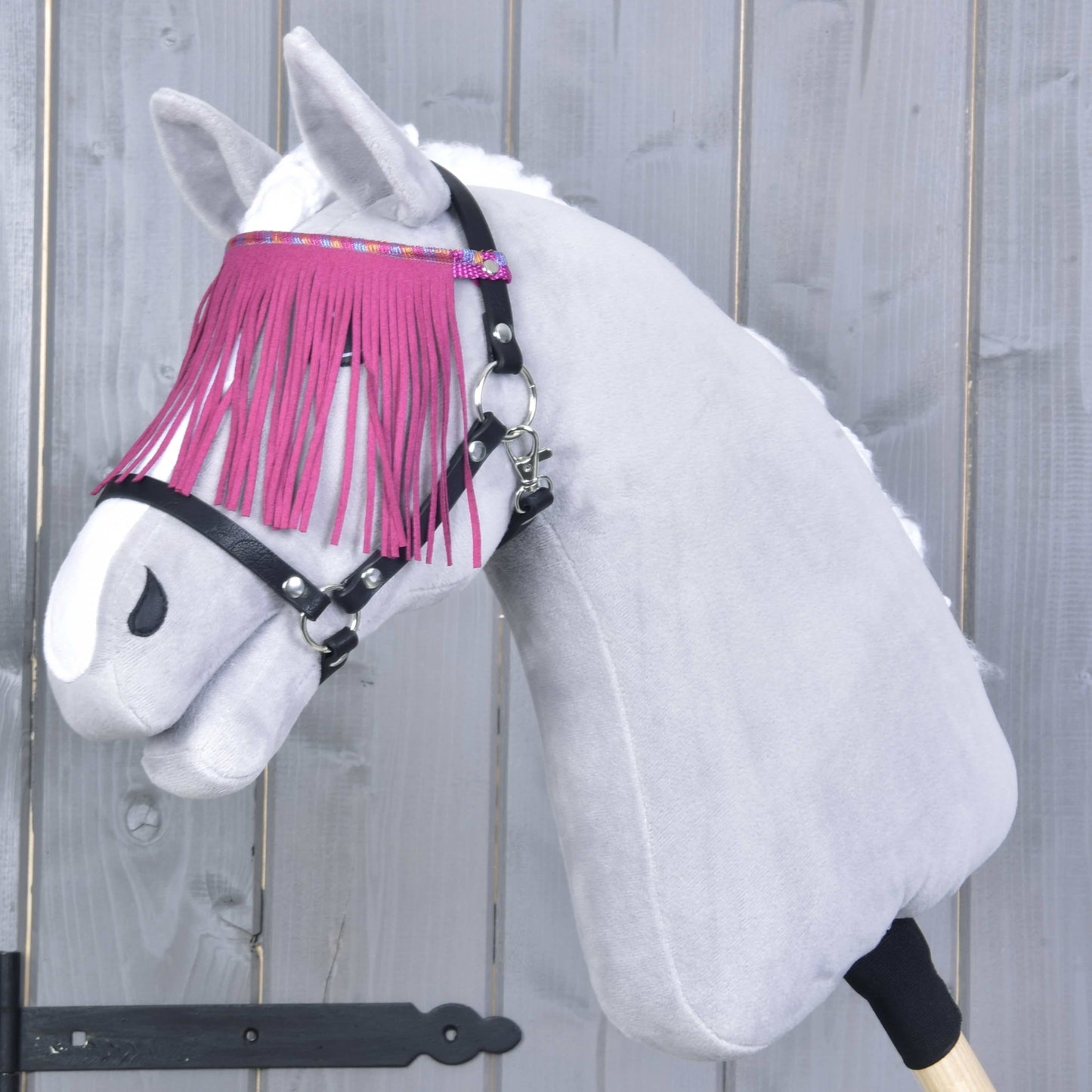 Hobby Horse en peluche de Hobby Horse LarDen portant le Licol noir avec franges roses, devant un fond en bois gris, avec robe blanche et têtière élégante à franges roses.