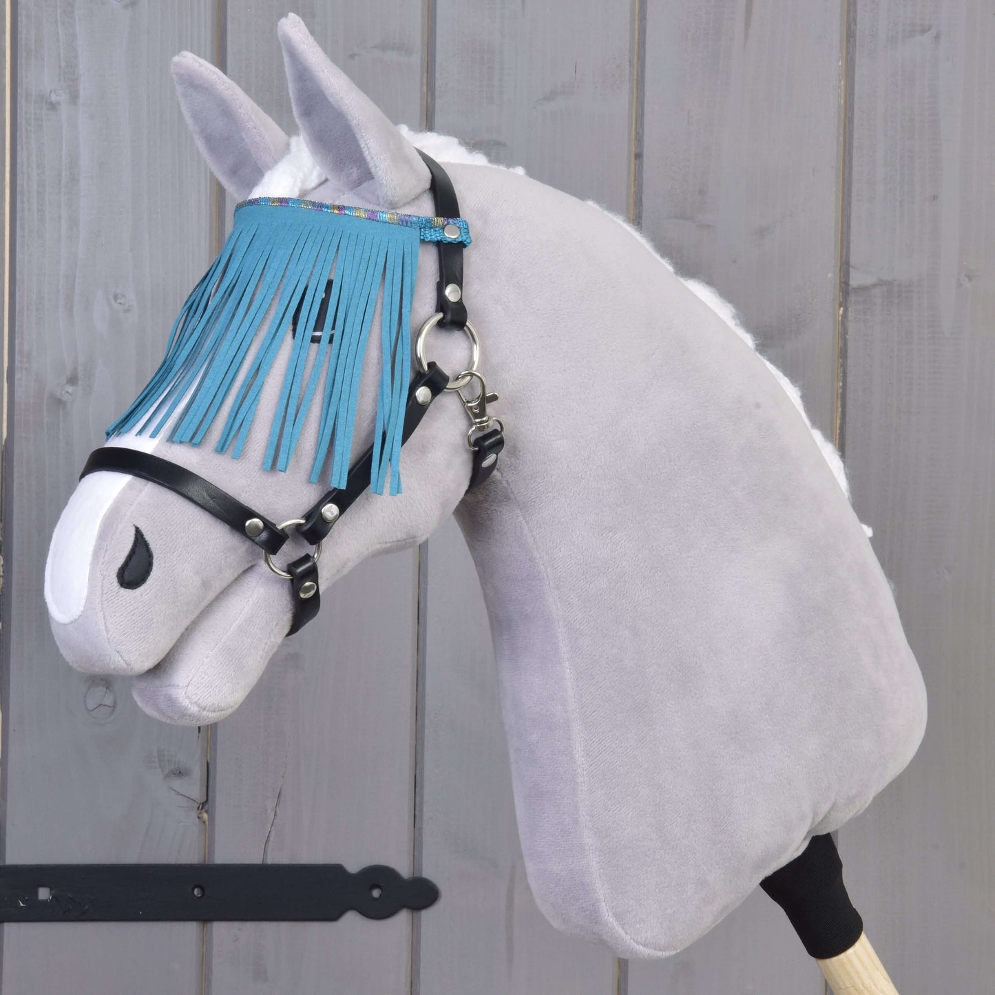 Hobby Horse LarDen gris avec licol noir à franges turquoise devant un mur en bois.