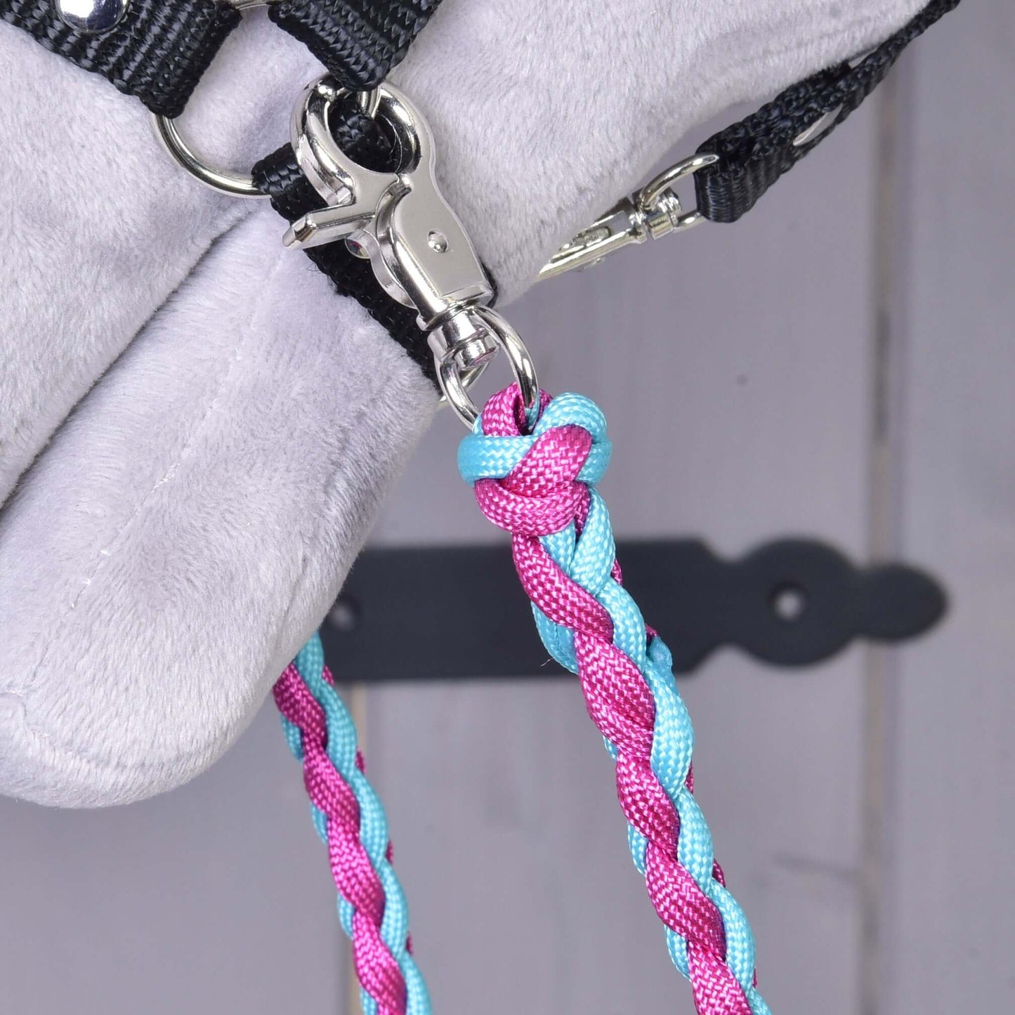 Gros plan sur les rênes tricotées rose et bleu de Hobby Horse LarDen, montrant un mousqueton métallique fixant les rênes en corde de polypropylène à une sangle noire sur une surface gris clair. Idéal comme configuration de rênes pour Hobby Horse. Porte en bois floue avec charnière noire en arrière-plan.