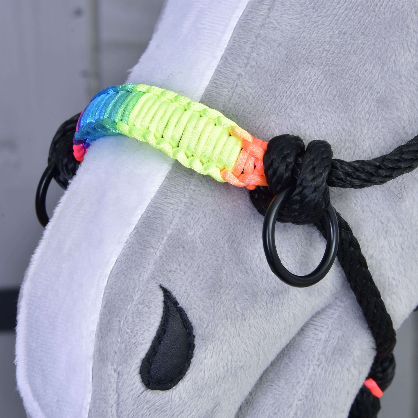 Gros plan d'un Hobby Horse gris en peluche de Hobby Horse LarDen, portant le Licou en corde Arc-en-ciel avec anneaux en métal et muserolle arc-en-ciel vive. Une marque noire en forme de goutte est visible sur son visage.