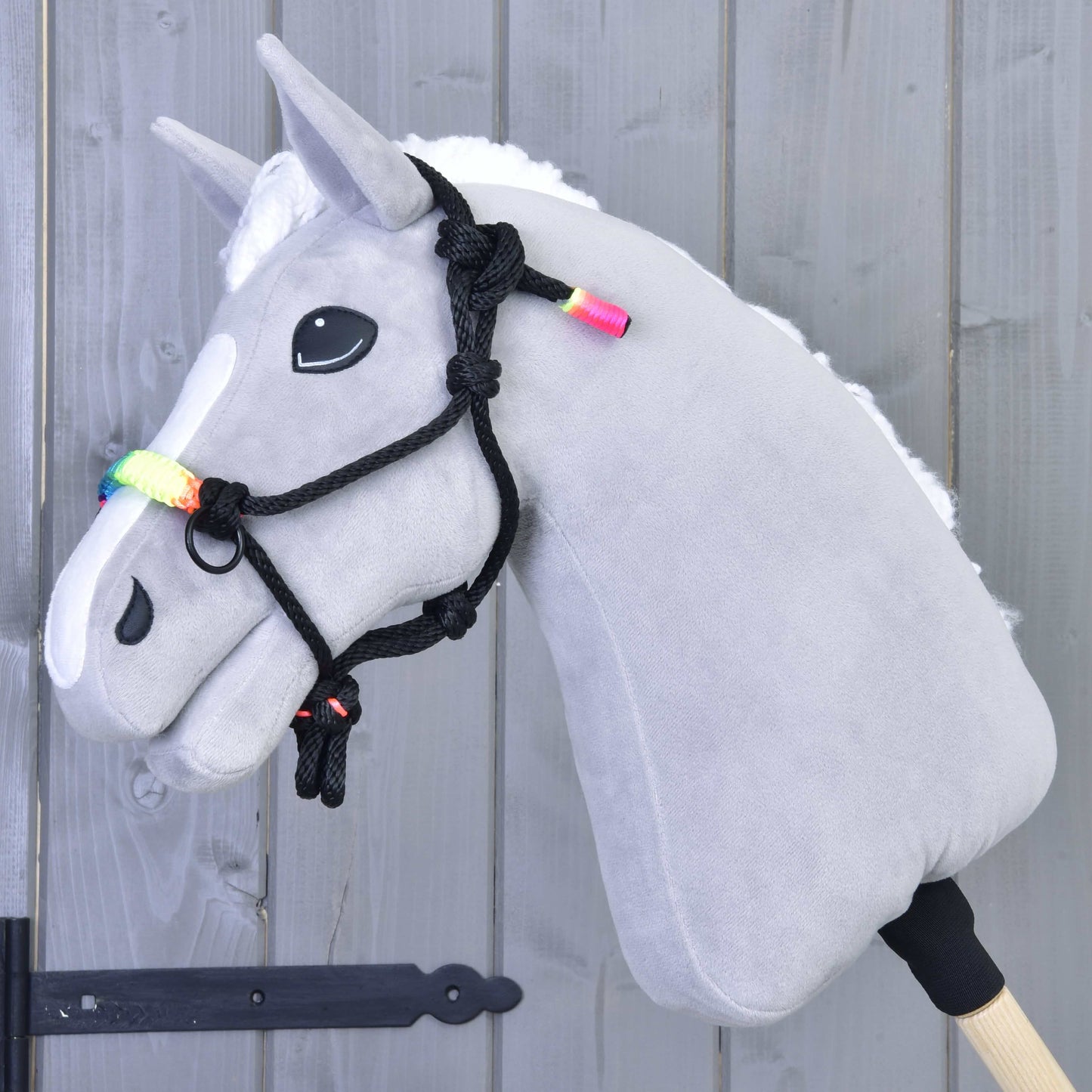 Licou en corde Arc-en-ciel de Hobby Horse LarDen, Hobby Horse gris avec crinière blanche, perles aux couleurs de l'arc-en-ciel et licou en corde noir sur un bâton en bois, présenté devant un mur en bois gris clair.