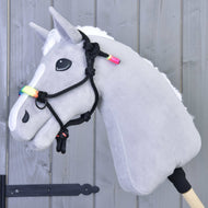 Hobby Horse LarDen Seilhalfter Regenbogen an einem grauen Hobby Horse mit weißer Mähne, regenbogenfarbenen Perlen und schwarzem Seilhalfter auf einem Holzstab, vor einer hellgrauen Holzwand.