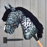 Hobby Horse Picasso mit schwarzem Halfter von Hobby Horse LarDen, gestaltet als markanter Hengst mit schwarz-weißem Leopardenmuster, das an das getupfte Fell eines Appaloosa erinnert, mit schwarzer Garnmähne und schwarzem Halfter, auf einem Holzstab vor einer grauen Holztür.