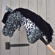 Hobby Horse Picasso mit schwarzem Halfter von Hobby Horse LarDen mit Plüschkopf in grau-schwarz gesprenkeltem, von einem Appaloosa inspiriertem Fellmuster, schwarzem Halfter und schwarzer Garnmähne, auf einem Holzstab vor einer grauen Holzwand.