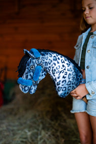 Mädchen in Jeansjacke hält das Hobby Horse Picasso mit schwarzem Halfter von Hobby Horse LarDen vor eine rote Scheunenwand aus Holz – blau-weißer Plüschkopf mit Leopardenmuster, schwarze Garnmähne – mit gefüttertem Teddy Petrol Halfter