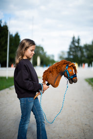 Lifestyle photo: a girl in a dark sweatshirt walks the Hobby Horse Pippa with Black Halter by Hobby Horse LarDen down a sandy track past the arena - chestnut plush head, ginger yarn mane - shown here with the fur-lined LarDen Blue halter and the Bloom Blue lead rope.