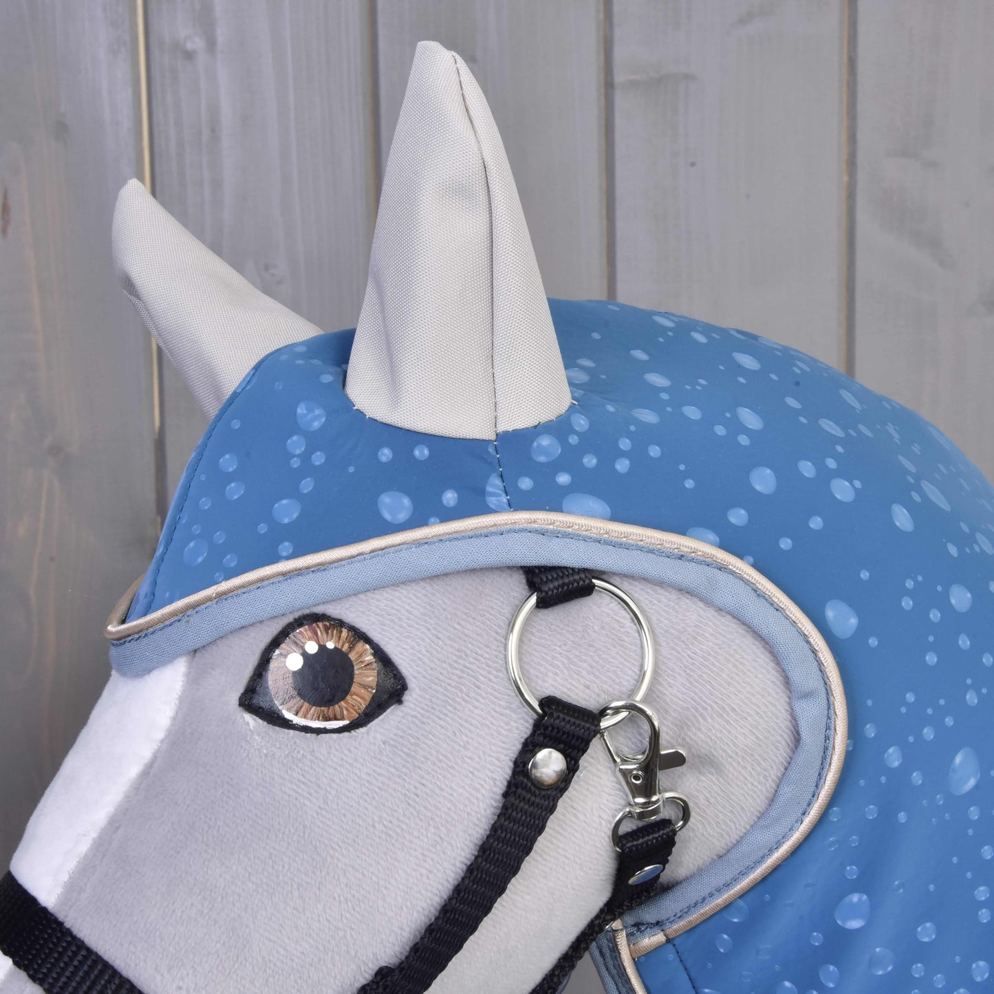 Nahaufnahme eines Kopfes mit Hobby Horse LarDen Regendecke Dolly (softshell) mit blauem Regenmantel mit Regentropfenmuster und schwarzem Halfter vor hellen Holzpaneelen – ideal als Spielzeug für jedes Wetter.